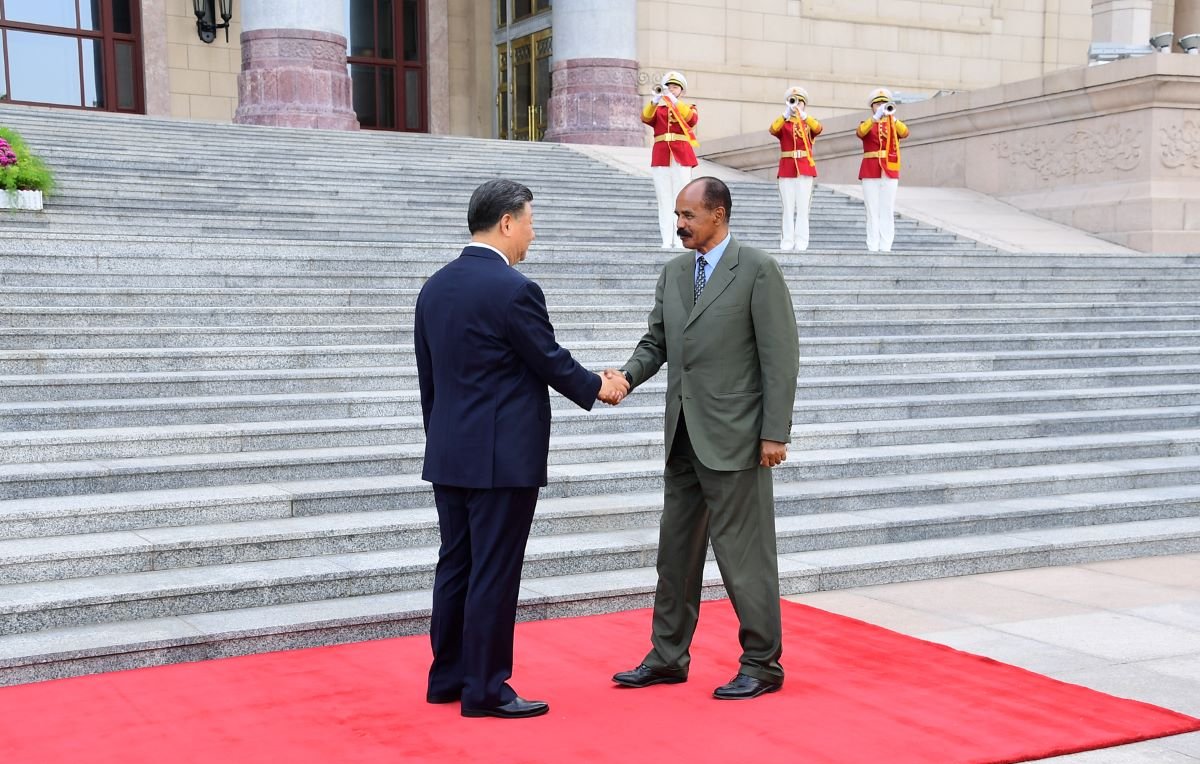 L'image montre deux hommes se serrant la main sur un tapis rouge, symbolisant un accueil officiel. Ils sont debout devant un bâtiment imposant, avec des marches qui mènent à l'entrée. En arrière-plan, on aperçoit des gardes en uniformes, debout et formant une ligne. Le premier homme porte un costume sombre, tandis que le second est habillé d'un costume vert. L'atmosphère dégage un sentiment de cérémonie et de respect mutuel.