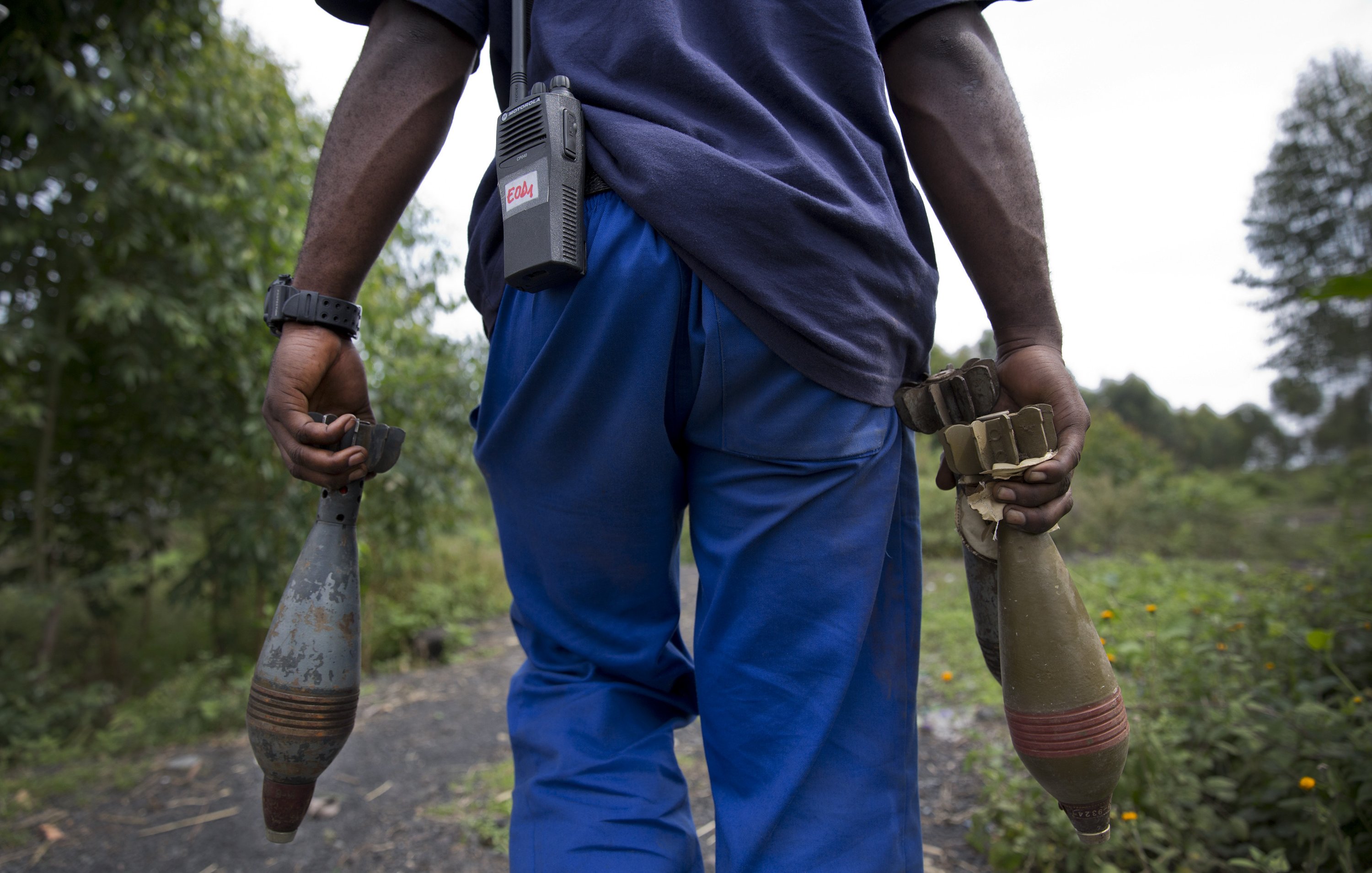 L'image montre un homme vu de dos, portant des vêtements décontractés : un t-shirt bleu foncé et un pantalon bleu. Il tient dans chaque main un objet long et cylindrique, probablement des munitions ou des dispositifs explosifs. À sa taille, il y a un appareil de communication. En arrière-plan, on aperçoit une nature verdoyante, avec des arbres et des plantes. L'ambiance semble à la fois sérieuse et chargée d'une importance qui nécessite prudence et vigilance.