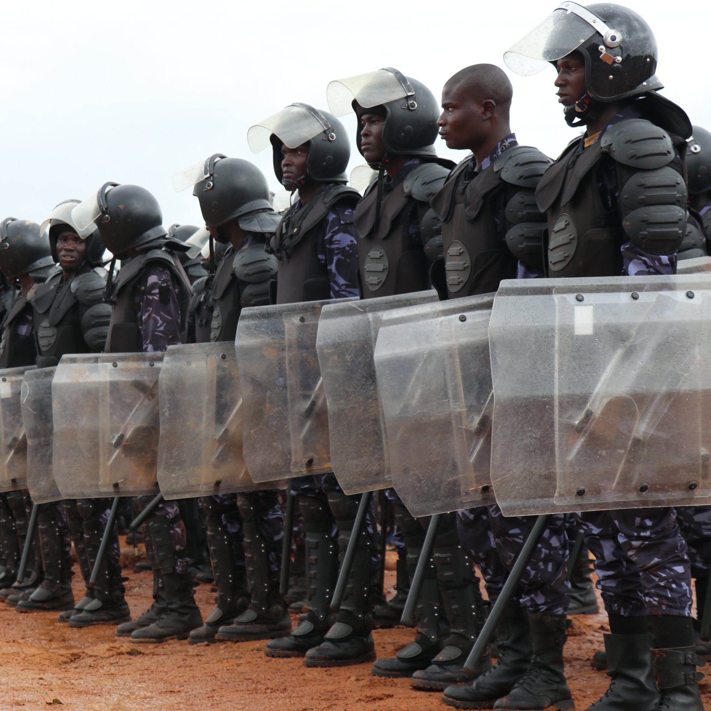 L'image montre une formation de policiers en tenue anti-émeute. Ils se tiennent en ligne, rigides et organisés, en portant des casques de protection et des gilets pare-balles. Chaque policier tient un bouclier transparent en face de lui, prêt à faire face à une situation potentielle. Le sol est en terre battue et l'environnement semble être à l'extérieur, avec quelques nuages visibles dans le ciel. Au loin, on peut apercevoir des personnes qui observaient la scène. L'atmosphère suggère une présence de sécurité renforcée.
