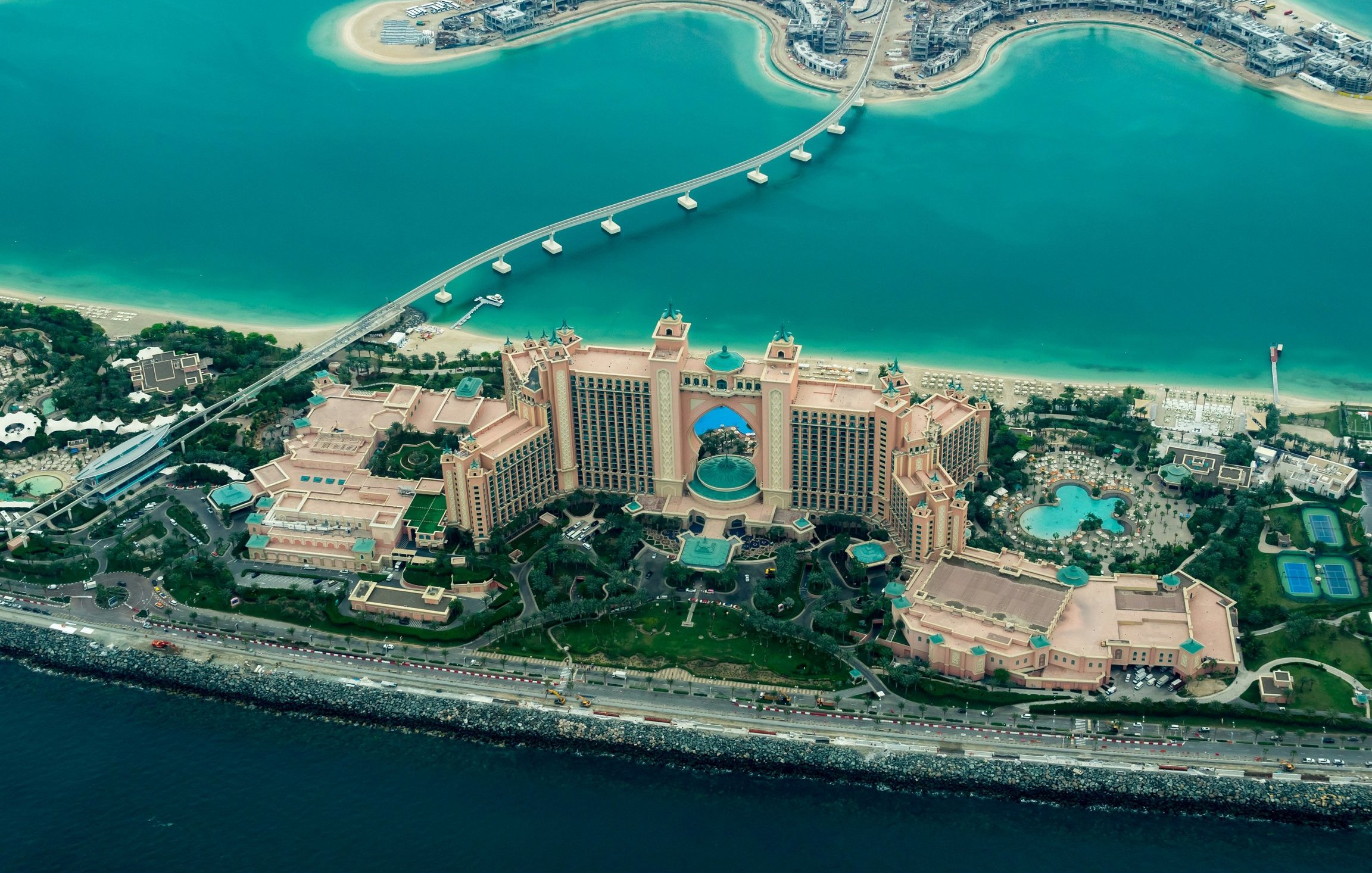 Cette image offre une vue aérienne d'un majestueux complexe hôtelier, entouré par des eaux turquoise. Le bâtiment principal, qui semble imposant, est décoré de nuances de beige et d'or, avec une architecture inspirée des contes arabes. Des jardins luxuriants entourent l'hôtel, agrémentés de piscines scintillantes et de palmiers. À proximité, on peut voir un chemin qui s'étend au-dessus de l'eau, reliant le terre-plein à d'autres structures. L'ensemble dégage une atmosphère luxueuse et tropicale, évoquant des vacances chaleureuses.