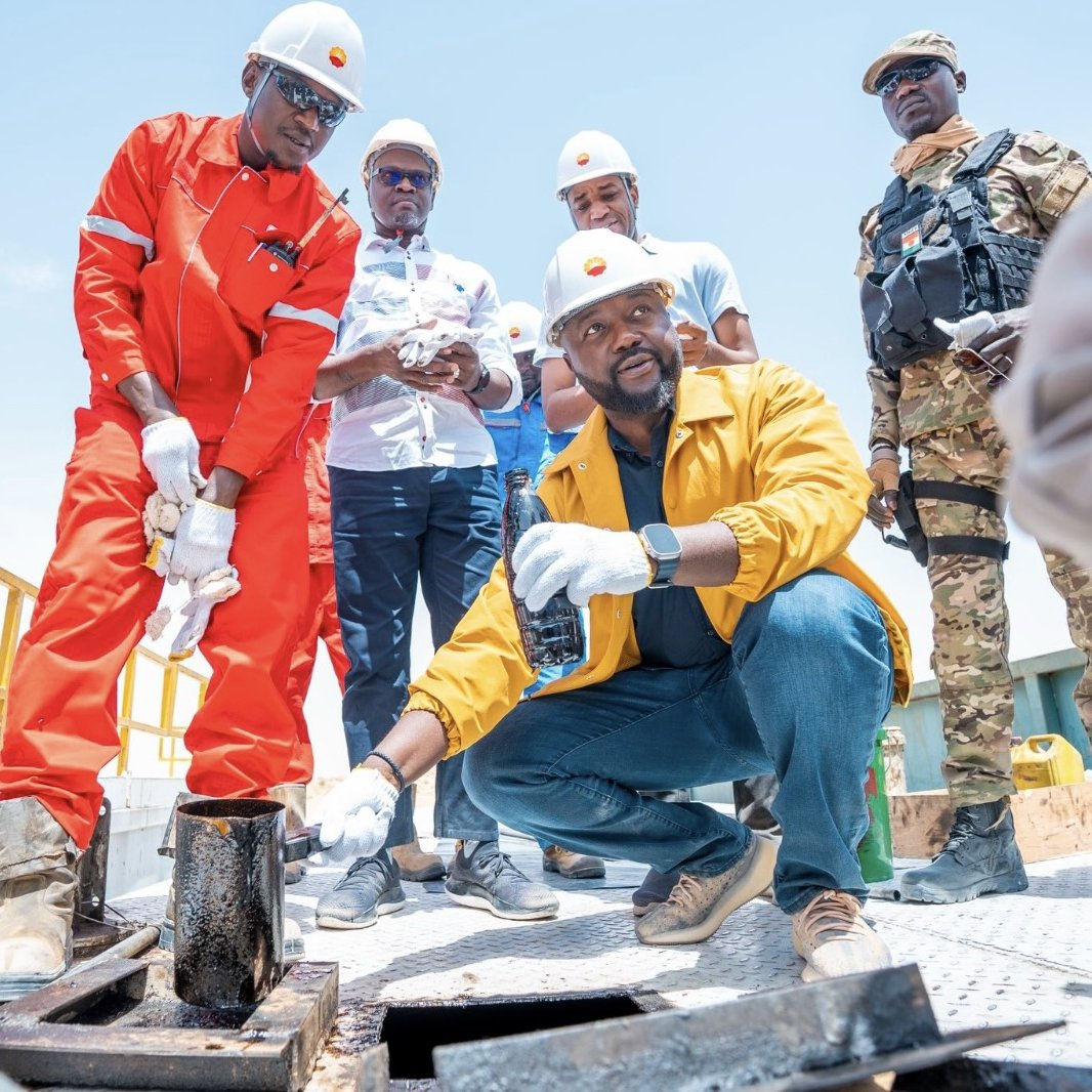 Dans cette image, on voit des hommes en tenue de travail sur une plateforme en extérieur. Deux d'entre eux portent des casques de sécurité blancs et des vêtements de protection, l'un en orange et l'autre en jaune. Le premier, en orange, se penche pour examiner un récipient, tandis que le second, en jaune, est à genoux et tient un échantillon de liquide dans une bouteille. En arrière-plan, d'autres personnes regardent, vêtues de manière décontractée. Le ciel est dégagé, suggérant une journée ensoleillée. L'environnement semble être lié à un travail industriel ou d'ingénierie, avec des installations visibles autour de eux.