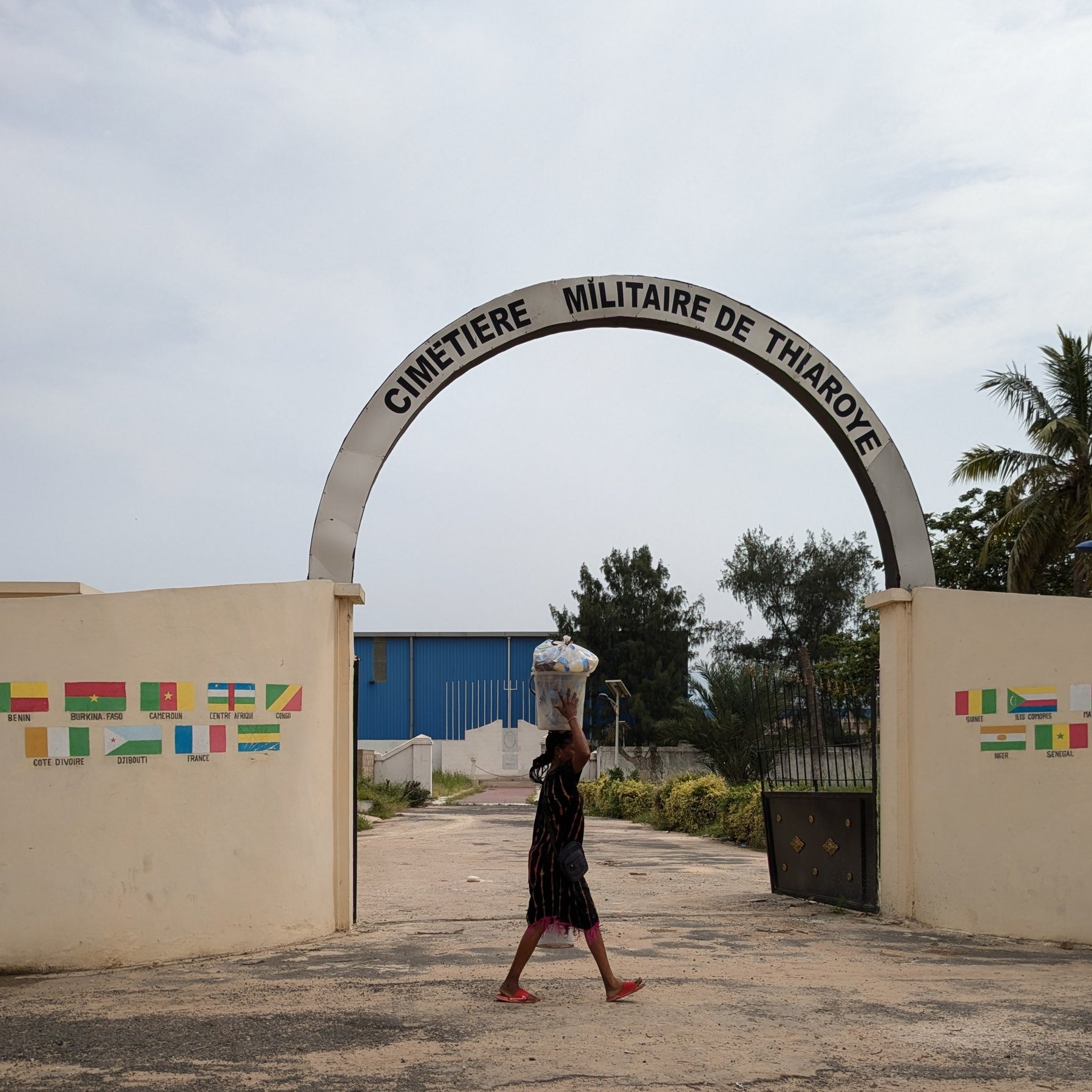 The image shows a person walking through the entrance of "Cimetière Militaire de Thiaroye," a military cemetery. The archway is prominently displayed, with the name of the cemetery written above. On either side of the entrance, there are colorful flags painted on the walls. The person in the foreground is carrying a large container on their head, and the scene is partly illuminated by natural light, surrounded by palm trees and greenery. The sky is overcast, giving a muted ambiance to the setting.