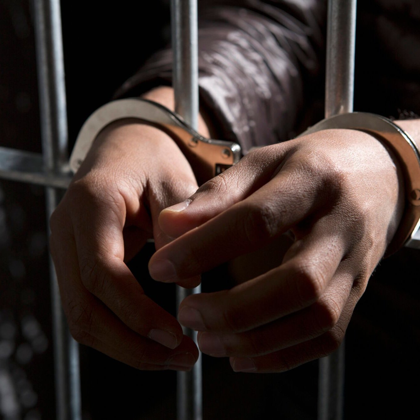 L'image montre des mains menottées, immobilisées par des menottes métalliques, qui se trouvent derrière des barreaux. Les mains sont tendues et légèrement fléchies, occupant une position qui évoque à la fois la vulnérabilité et la contrainte. Le fond est sombre, créant un contraste avec la lumière qui met en évidence les éléments métalliques des menottes et les détails de la peau. L'ensemble de la scène transmet une ambiance de confinement et de détention.