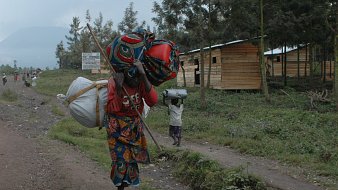 L'image montre un paysage rural d'une région montagneuse, probablement en Afrique. Sur la droite, vous voyez des maisons en bois, simples et fonctionnelles, avec un accent sur la nature environnante. À l'avant, une femme porte une charge lourde sur son dos, maintenue par un tissu coloré. Elle est vêtue d'une robe traditionnelle aux motifs vifs et colorés, qui contrastent avec le sol terreux et les feuillages verts. Dans le fond, une autre personne transporte également une charge, symbolisant la dureté du travail dans cette communauté. Les montagnes se dessinent en arrière-plan, offrant un sentiment d'immensité et de tranquillité, tandis que la scène évoque la résilience et le quotidien des habitants de cette région.