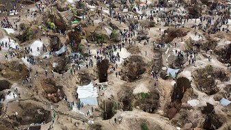 L'image représente une grande site d'extraction minière, probablement un lieu de prospection. On y voit de nombreuses personnes, qui semblent être des mineurs ou des chercheurs de minerai, dispersées sur un terrain accidenté. Le sol est parsemé de nombreuses cavités, témoignant des efforts de creusement. Certaines zones sont abritées par des abris temporaires en toile, tandis que d'autres sont à ciel ouvert. La scène dégage une dynamique de travail acharné, où les individus s'affairent dans un environnement à la fois désordonné et organisé, caractérisé par des monticules de terre et des roches. L'ensemble évoque un lieu de quête et de lutte pour des ressources précieuses.