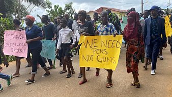 Dans cette image, un groupe de personnes marche ensemble dans une rue. On peut entendre des voix et des chants de solidarité. Les participants tiennent des pancartes aux couleurs vives. Certaines pancartes affichent des messages puissants comme "WE NEED PENS NOT GUNS", exprimant une demande pour la paix et l'éducation plutôt que pour la violence. Les manifestants sont vêtus de différents styles de vêtements, allant de tenues formelles à des vêtements décontractés. En arrière-plan, on aperçoit des maisons et des arbres qui ajoutent un cadre naturel à cette scène de protestation pacifique. L'atmosphère est déterminée et engagée, reflet d'un désir de changement et d'un message fort pour la communauté.