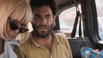 Dans cette image, nous sommes dans l'intérieur d'un véhicule, probablement un taxi ou une voiture. À l'avant, deux personnes sont assises. À gauche, une femme, dont les cheveux sont très blonds et coupés court, porte des lunettes de soleil. Elle semble attentive et se penche un peu vers la droite. À droite, un homme avec une barbe et des cheveux frisés regarde devant lui, semblant préoccupé ou surpris. L'ambiance semble tendue, et l'intérieur de la voiture est éclairé par la lumière extérieure. On peut également apercevoir d'autres véhicules à l'extérieur par les fenêtres.