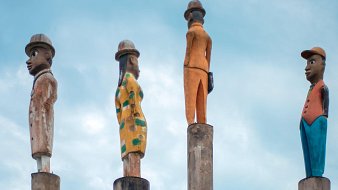 L'image présente quatre sculptures stylisées sur des colonnes. Chaque sculpture est peinte de manière différente, représentant des personnages humains : 1. La première sculpture à gauche est un homme avec un chapeau, vêtu d'un costume. Son expression est sérieuse. 2. La deuxième sculpture est une femme qui porte une robe avec un motif à rayures. Elle se tient droite, les bras le long du corps. 3. La troisième sculpture est un homme en orange, qui se tient également droit mais légèrement tourné sur le côté. 4. Enfin, la dernière sculpture est un homme en costume bleu, avec un chapeau, regardant vers l'horizon. L'arrière-plan est dominé par un ciel bleu clair, créant une ambiance paisible. Les sculptures semblent faire partie d'une installation artistique qui évoque la culture et l'identité. Les colonnes sur lesquelles elles reposent ajoutent une touche de hauteur et de grandeur à la scène.