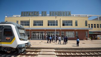 The image shows the exterior of a train station labeled "ESTAÇÃO DE BENGUELA." The building has a modern architectural design with large glass windows and a clean facade. In front of the station, a group of people, likely staff or passengers, are gathered. To the left, there is a sleek train parked on the tracks, showcasing a contemporary design. The scene is bright, indicating a clear blue sky. The overall setting suggests a bustling transportation hub.