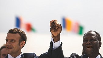 The image shows two men, one wearing a suit with a tie, raising their hands together in a gesture of solidarity or celebration. They are smiling and appear to be in a public setting. In the background, there are several flags visible, suggesting an international context or event. The atmosphere seems positive and hopeful.