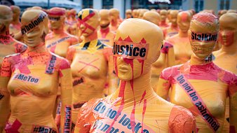 L'image montre une installation artistique composée de nombreux mannequins recouverts de bandes de plastique colorées, principalement dans des teintes orange et jaune. Ces mannequins sont alignés et se font face, créant une impression de masse. Sur leur surface, on peut voir des mots et des phrases comme "justice", "Stalking", "MeToo" et "Do not look away", écrits en noir et rouge, ce qui suggère un message fort et engagé. L'ambiance générale semble à la fois frappante et troublante, évoquant des thèmes de lutte contre l'injustice et les violences faites aux femmes. Les couleurs vives et les slogans bien en vue attirent l'attention et suscitent réflexion.