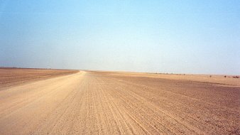 The image depicts a barren landscape characterized by a vast, empty stretch of land, likely a desert or arid region. The foreground features a dirt road that leads into the distance, flanked by dry, flat terrain. The sky overhead is clear and blue, creating a stark contrast with the earthy tones of the ground. The overall scene conveys a sense of isolation and openness, typical of remote, dry environments.