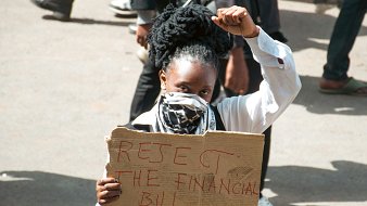 The image depicts a protester holding a cardboard sign that reads "REJECT THE FINANCIAL BILL PUNNYASS." The individual is wearing a white shirt and has their hair styled in large, coiled forms. They are raising a fist in a gesture of defiance or solidarity, and are also wearing a mask over their face. The background includes other people and a busy street scene, suggesting a gathering or demonstration.