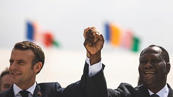 Imaginez une scène marquée par un moment de camaraderie et d'unité. Deux hommes se tiennent côte à côte, levant la main droite en l'air, leurs poings serrés, symbolisant la force et la solidarité. À gauche, on perçoit un homme avec une coupe de cheveux soignée, vêtu d'un costume sombre, tandis que l'homme à droite, également en costume, affiche un grand sourire chaleureux. En arrière-plan, plusieurs drapeaux flottent doucement au vent, représentant différentes nations. L'atmosphère est festive et pleine d'espoir.