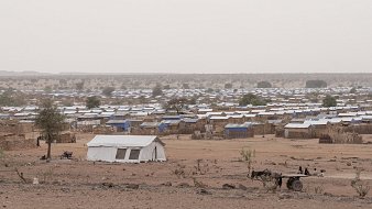 L'image montre un camp de réfugiés. Au premier plan, un enfant assis sur un amas de coussins et de matelas, portant un t-shirt rouge. Il semble pensif, regardant au loin. À côté de lui, un autre enfant est assis sur une petite table, vêtu d'un t-shirt bleu. L'environnement est poussiéreux, avec plusieurs tentes blanches en arrière-plan. On peut également apercevoir des objets éparpillés, comme des cartons et des bidons, évoquant une vie temporaire et difficile. L'atmosphère semble mélancolique et montre une réalité de vulnérabilité.