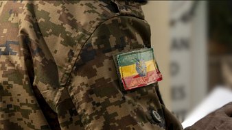 The image features a close-up of a military uniform with a camouflage pattern. Prominently displayed on the arm is a patch showing the flag of Ethiopia, which consists of horizontal stripes in green, yellow, and red, with a blue circle and a yellow star in the center. The fabric of the patch appears worn, adding to its character.