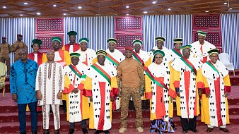 The image features a formal gathering of a group of individuals, likely in a ceremonial setting. They are wearing traditional attire, which includes colorful robes adorned with striped and patterned elements. Many of them are also wearing distinctive hats or headgear. The setting appears to be well-decorated, possibly for an official event, with a red carpet and banners or decorations in the background. A few individuals stand out in military or official uniforms, suggesting a mix of civilian and military presence at the event.