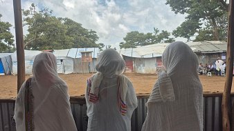 L'image montre trois femmes de dos, se tenant côte à côte, regardant à travers une structure en bois ouverte. Elles portent des vêtements longs et légers. Leurs têtes sont couvertes de foulards, ce qui suggère un contexte culturel ou religieux. Au loin, on aperçoit des abris temporaires, probablement des tentes ou des structures en tôles. Le ciel est partiellement nuageux, créant une atmosphère paisible. L'environnement évoque un lieu de rassemblement ou d'attente, avec un sol en terre battue. La scène semble calme, laissant entrevoir un moment de contemplation ou de partage entre ces femmes.