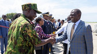 The image shows a formal scene at an airport where a group of people is gathered, likely to welcome a dignitary or a visitor. In the foreground, two men are shaking hands; one is dressed in military camouflage, and the other is in a formal suit. Around them, there are officials in uniform and business attire lined up, indicating a significant or ceremonial occasion. The setting seems to be sunny, contributing to a positive atmosphere.