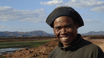 The image features a smiling man wearing a dark hat and a sweater. He is standing in an outdoor setting, likely near agricultural land, with a backdrop of hills and a partly cloudy sky. The ground appears to be bare earth, suggesting possible farming activity nearby. His cheerful expression conveys a sense of warmth and friendliness.