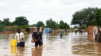 The image depicts a flooded area, with individuals wading through water that has inundated the surroundings. The water reaches knee-deep, affecting homes and structures in the background, which appear to be partially submerged. People in the scene are carrying various items, suggesting they're either evacuating or salvaging belongings. Trees and greenery can be seen around the area, indicating that it is a rural or semi-urban environment. The sky is overcast, contributing to the somber atmosphere of the flooding situation.