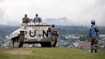 The image depicts a scene from a military operation involving United Nations peacekeepers. In the foreground, a military vehicle marked with "UN" is parked on a grassy area overlooking a landscape. Several soldiers in blue helmets, which signify their affiliation with the UN, are positioned around the vehicle. They appear to be surveying the area, which includes a view of a village with rooftops in the background. The atmosphere seems calm, with a cloudy sky adding to the setting. The soldiers' uniforms suggest they are part of a peacekeeping mission, emphasizing their role in maintaining stability in the region.