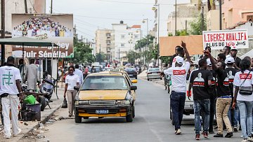 L'image montre une scène animée dans une rue urbaine. À gauche, des personnes marchent, certaines portant des tee-shirts blancs avec des inscriptions visibles. Ils semblent participer à une manifestation ou un rassemblement. Au centre, un taxi jaune est garé sur le côté de la route. Sur le côté droit, il y a plusieurs personnes, et l'ambiance donne une impression de dynamisme et d'engagement. Des affiches sont visibles en arrière-plan, dont l'une mentionne des slogans politiques, suggérant un contexte de revendication ou de mobilisation sociale. Le bruit de la ville, les voix des manifestants et le bruit des véhicules complètent cette atmosphère vivante.