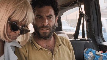 Dans cette image, nous sommes dans l'intérieur d'un véhicule, probablement un taxi ou une voiture. À l'avant, deux personnes sont assises. À gauche, une femme, dont les cheveux sont très blonds et coupés court, porte des lunettes de soleil. Elle semble attentive et se penche un peu vers la droite. À droite, un homme avec une barbe et des cheveux frisés regarde devant lui, semblant préoccupé ou surpris. L'ambiance semble tendue, et l'intérieur de la voiture est éclairé par la lumière extérieure. On peut également apercevoir d'autres véhicules à l'extérieur par les fenêtres.