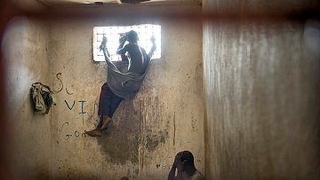 L'image dépeint un environnement sombre et restreint, vraisemblablement une cellule ou une pièce fermée. Au premier plan, deux personnes sont présentes. L'une est assise, visiblement accroupie, la tête entre les mains, adoptant une posture de détresse ou de réflexion. L'autre personne est perchée près d'une fenêtre grillagée, les jambes repliées, enveloppée dans un drap qui semble être une couverture. La lumière qui pénètre par la fenêtre est limitée, créant un contraste entre l'extérieur et l'intérieur sombre de la pièce. Les murs portent des marques, probablement des graffiti, ce qui suggère un environnement négligé. L'atmosphère générale est lourde et empreinte de mélancolie, reflétant un sentiment d'isolement et de désespoir.