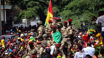 L'image montre une scène de célébration animée. Au premier plan, un groupe de soldats en uniforme militaire se tient debout, certains brandissant un drapeau aux couleurs vives de la Guinée, rouge, jaune et vert. Ils semblent être au centre d'une foule massive, qui est remplie de personnes qui portent également des vêtements aux couleurs du drapeau. L'ambiance est festive et euphorique, avec des visages souriants et levés vers les soldats. En arrière-plan, on aperçoit des arbres et des bâtiments, créant un cadre urbain. L'énergie collective de la foule et la présence militaire suggèrent un événement significatif, probablement une célébration ou une manifestation patriotique.