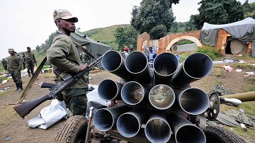 L'image montre un soldat en uniforme, debout à côté d'un véhicule équipé de plusieurs tubes visibles en rangée, qui semblent être des lance-roquettes. Le soldat tient une arme à feu, se tenant avec sérieux. En arrière-plan, on aperçoit d'autres soldats, ainsi qu'un environnement chaotique où des objets sont éparpillés, ce qui donne l'impression d'une zone de conflit ou de préparation militaire. Le tout se déroule dans un paysage verdoyant, avec des collines au loin.