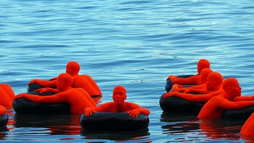 Cette image présente un groupe de sculptures humaines d'une couleur orange vif, flottant sur une surface d'eau calme. Chaque sculpture est assise ou allongée sur des anneaux flottants noirs, composant un contraste frappant avec le bleu azur de l'eau. Les sculptures ont des formes simplifiées, sans détails distincts de visage ou de vêtements, ce qui les rend abstraites. L'ensemble dégage une atmosphère à la fois sereine et intrigante, où la composition des éléments et les couleurs vives attirent l'attention.