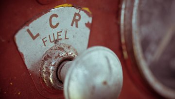 L'image montre un gros plan d'un ancien indicateur de carburant sur une machine, probablement un tracteur ou un véhicule vintage. La partie visible présente un disque circulaire marqué avec les lettres "LCR" et le mot "FUEL" en dessous. La surface est usée, avec des éraflures et une couleur rouge qui témoigne de son âge. À côté, il y a un bouton en métal brillant, légèrement terni, qui ajoute à l'aspect nostalgique de l'objet. L'ensemble évoque une impression de robustesse et d'histoire, typique des équipements anciens.