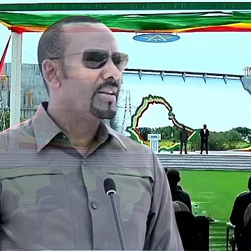 L'image montre un homme debout au premier plan, portant des lunettes de soleil et un uniforme militaire, qui semble s'adresser à un public. Il est probablement en train de faire un discours. En arrière-plan, on aperçoit une grande scène décorée de drapeaux éthiopiens et d'autres couleurs vives, avec une grande structure ressemblant à un barrage. Au premier plan, on peut voir des personnes assises, attentives, apprêtées pour un événement officiel en plein air, sur une pelouse verte. L'atmosphère semble d'une grande importance, peut-être liée à une célébration ou une inauguration.