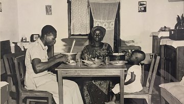 L'image en noir et blanc montre une scène de repas familial dans une pièce simple, probablement une cuisine. À une table en bois, un homme en uniforme, assis sur une chaise, semble être en train de manger avec une grande cuillère. En face de lui, une femme, vêtue d'une robe traditionnelle et d'un chapeau, regarde avec un sourire bienveillant. À côté d'elle, un petit enfant est assis sur une autre chaise, portant une petite robe blanche, observant la scène. Les murs de la pièce sont décorés sobrement, et il y a des verres sur la table, ajoutant une touche de quotidien à ce moment plein de convivialité.