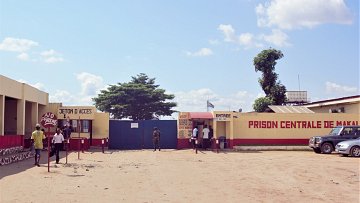 The image depicts the entrance to a central prison, labeled "PRISON CENTRALE DE MAKALA." The scene shows a sandy area with a few people walking near the entrance. On the left, there is a booth or kiosk marked "JETON D'ACCÈS," and on the right, another booth labeled "ENTRÉE." In the background, there are trees and a clear blue sky with some clouds. A few vehicles are parked nearby, adding to the overall setting of the prison facility.