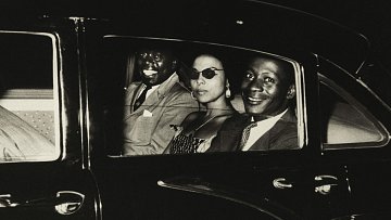 L'image présente trois personnes assises à l'intérieur d'une voiture sombre. L'ambiance semble élégante et vintage. À gauche, un homme souriant, au teint foncé, porte un costume clair. Au centre, une femme avec des lunettes de soleil stylées, habillée d'une robe à motifs, dégage une aura de sophistication. À droite, un autre homme, également souriant, avec un teint légèrement plus clair, porte une tenue qui semble chic. Les visages des occupants sont mis en valeur par un éclairage doux, créant une atmosphère chaleureuse et amicale. Le tout suggère un moment de convivialité, peut-être après une sortie ou un événement.