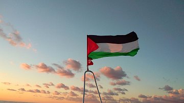 L'image montre un drapeau palestinien flottant sur un mât, avec un vaste horizon marin au fond. Le ciel est teinté de couleurs chaudes, probablement à l'aube ou au crépuscule, avec des nuages épars. Les couleurs du drapeau sont distinctes : le noir, le blanc, le vert et le rouge. La mer est calme et reflète doucement les teintes du ciel, créant une ambiance paisible et solennelle.