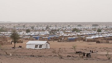 L'image montre un camp de réfugiés. Au premier plan, un enfant assis sur un amas de coussins et de matelas, portant un t-shirt rouge. Il semble pensif, regardant au loin. À côté de lui, un autre enfant est assis sur une petite table, vêtu d'un t-shirt bleu. L'environnement est poussiéreux, avec plusieurs tentes blanches en arrière-plan. On peut également apercevoir des objets éparpillés, comme des cartons et des bidons, évoquant une vie temporaire et difficile. L'atmosphère semble mélancolique et montre une réalité de vulnérabilité.