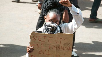 L'image montre une manifestation dans laquelle une femme se tient au premier plan. Elle a des cheveux noirs et crépus coiffés en chignon. Elle porte un masque sur le visage et un haut blanc. Dans sa main, elle élève le poing, symbole de résistance. L'autre main tient une pancarte en carton sur laquelle sont inscrits en lettres rouges les mots "REJECT THE FINANCIAL BILL PUNNYASS". En arrière-plan, on distingue d'autres personnes, soulignant le caractère collectif de la protestation. L'atmosphère est chargée d'énergie, avec un sentiment d'engagement palpable.