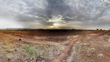 Imaginez un vaste paysage ouvert, dominé par des tons chauds de terre et des nuances de beige. Le sol est sec et parsemé de petites pierres, avec des herbes jaunies qui émergent de la terre aride. À l'horizon, un ciel nuageux s'étend, teinté de gris avec des rayons de lumière qui percent, annonçant un coucher de soleil. La lumière crépusculaire crée une ambiance paisible, tandis que la nature environnante semble calme et tranquille. Les silhouettes lointaines de collines ajoutent une dimension au paysage, renforçant le sentiment d'immensité et d'isolation.