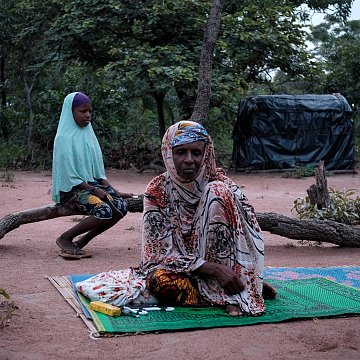Dans cette image, nous sommes dans un environnement naturel, avec le sol recouvert de terre rouge et entouré d'arbres. À l'avant, une femme est assise sur un tapis, vêtue d'un habit coloré et d'un foulard. Elle a une expression calme. À l'arrière-plan, une jeune fille est assise sur une branche, portant un voile et un vêtement aux motifs colorés. La scène dégage une atmosphère de sérénité et de contemplation, avec la lumière naturelle qui crée une ambiance douce et paisible.