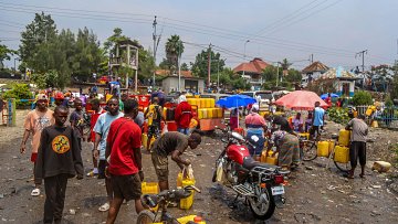 L'image représente une scène animée dans un quartier urbain. On peut imaginer un marché ou un point de distribution d'eau, où de nombreuses personnes sont rassemblées. Des hommes et des femmes, de divers âges, s'affairent autour de grands récipients jaunes qu'ils remplissent d'eau. Le sol semble en désordre, avec des déchets visibles, ajoutant une impression de négligence dans l'environnement. Des parapluies colorés sont déployés, fournissant un peu d'ombre sous un ciel légèrement nuageux. Au fond, on peut apercevoir des bâtiments et des arbres, tandis qu'un motard fait la queue avec sa moto, indiquant une activité routière environnante. L'atmosphère est dynamique, avec une forte interaction entre les gens.