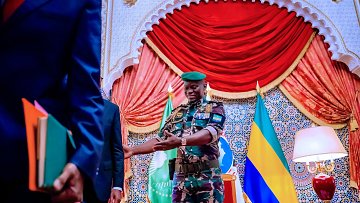 L'image montre un salon d'apparat coloré, avec un décor richement orné et des drapeaux en arrière-plan, dont on distingue le drapeau du Gabon. Au centre, un homme portant un uniforme militaire camouflé et une casquette verte se tient debout avec un sourire accueillant. Il est en train de tendre la main vers une autre personne, probablement pour une poignée de main. Les murs sont décorés de motifs arabes et la scène dégage une atmosphère de formalité et de respect. Une lampe est placée à droite, ajoutant une touche de chaleur à l'environnement.