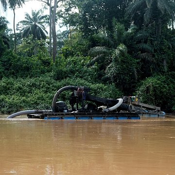 L'image montre un bateau sur une rivière aux eaux brunes. Ce bateau est équipé d'un grand tuyau qui semble aspirer de la matière de l'eau. À l'arrière-plan, on peut voir une végétation dense, composée de grandes plantes et d'arbres tropicaux, qui créent une ambiance naturelle et verdoyante. La scène évoque une activité liée à l'extraction de ressources, probablement de l'or ou des minéraux, dans un environnement de jungle.