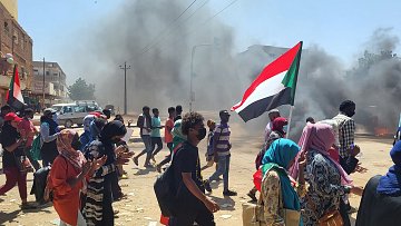 L'image montre une scène de manifestation animée. Des groupes de personnes marchent dans une rue, portant des drapeaux, dont un qui représente le Soudan. La foule paraît déterminée, et beaucoup de manifestants portent des masques, probablement en raison des conditions de pollution ou pour des raisons de sécurité. En arrière-plan, on peut voir des colonnes de fumée noire qui s'élèvent dans le ciel, créant une atmosphère tendue. Les manifestants semblent traverser une intersection, avec des véhicules garés sur le côté. L'atmosphère générale dégage une impression de forte mobilisation et de passion collective.
