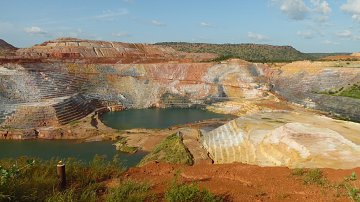 L'image montre une carrière de minerai, un paysage dramatique où les strates de terre créent des dégradés de couleurs allant du rouge au jaune et au blanc. Les parois de la carrière sont escarpées, formant des niveaux bien distincts. Au centre, un plan d'eau bleu émerge, contrastant avec les teintes terreuses et les paysages environnants. La végétation se fait rare sur les bords, mais quelques petites herbes vertes remontent le long des pentes. Le ciel est clair avec quelques nuages épars, ajoutant une lumière douce à la scène. Cette vue évoque à la fois l'exploitation humaine et la beauté naturelle brute.