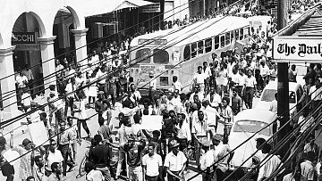 L'image que vous avez devant vous représente une scène urbaine animée dans les années 1960. On peut voir une foule dense de personnes marchant dans une rue. Les gens portent des vêtements typiques de l'époque, et il semble qu'ils se déplacent avec un but précis. À gauche, plusieurs commerces sont visibles, avec des enseignes en lettres visibles. Un ancien bus à l'allure caractéristique est garé, ajoutant une touche nostalgique à la scène. On aperçoit également quelques voitures stationnées et d'autres qui circulent dans la rue. Les fils électriques sont visibles en haut de l'image, accentuant l'impression d'une ville vivante et dynamique. L'atmosphère générale évoque une période de mouvement socioculturel important, avec des échanges et une vitalité palpable dans l'air.