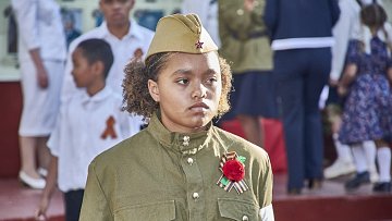 The image features a young girl wearing a military-style uniform with a khaki color. She has a military hat adorned with a star. The uniform includes decorative items like a red flower and a ribbon, possibly indicating a celebration or commemorative event. In the background, there are other children and adults dressed in formal attire, suggesting a public gathering or ceremony, likely related to a historical event. The setting appears to be outdoors, with a backdrop of steps and other participants.