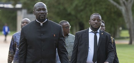 L'image montre un groupe d'hommes marchant ensemble sur un chemin dans un environnement verdoyant. Au premier plan, un homme grand et imposant porte un costume noir avec un col Mao, tandis qu'un autre homme à côté de lui est en costume noir et cravate. Ils ont l'air sérieux et concentrés, comme s'ils se rendaient à un événement important. En arrière-plan, d'autres hommes sont visibles, avec des tenues variées, ce qui suggère une atmosphère de réunion ou de conférence. Le paysage est paisible, avec des arbres et une lumière douce, ajoutant à l'ambiance formelle de la scène.